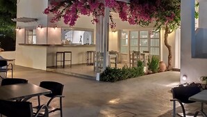 Un área de comedor al aire libre con mesas y sillas, una barra y un edificio de fachada blanca y plantas con flores rosadas.