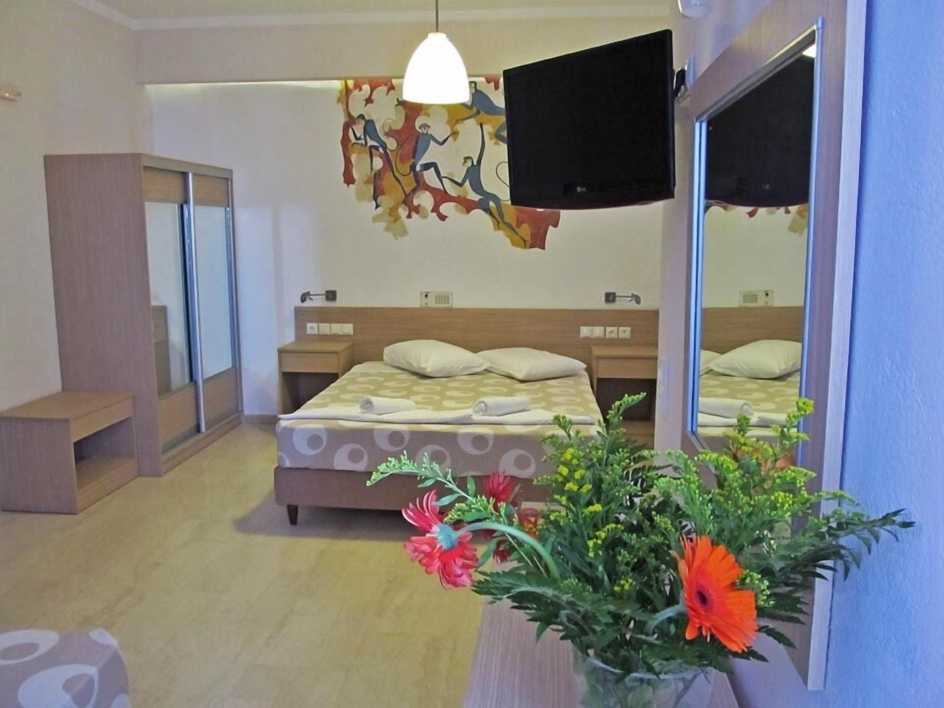 A hotel room with a bed, bedside tables, a TV, a mirror, and a flower arrangement.
