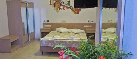 A hotel room with a bed, bedside tables, a TV, a mirror, and a flower arrangement.