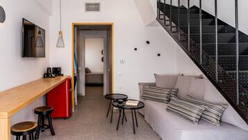 A modern living space with a wooden desk, two stools, a small round table, a sofa with striped pillows, and a staircase with a metal railing.