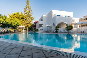 Una piscina con sillones de playa y sombrillas, rodeada de edificios blancos con detalles azules y abundante vegetación.