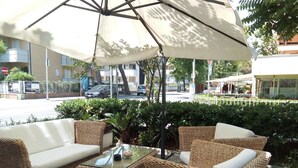 An outdoor seating area with wicker furniture and a glass-top table, shaded by a large umbrella. - Hotel Majorca (Riccione)