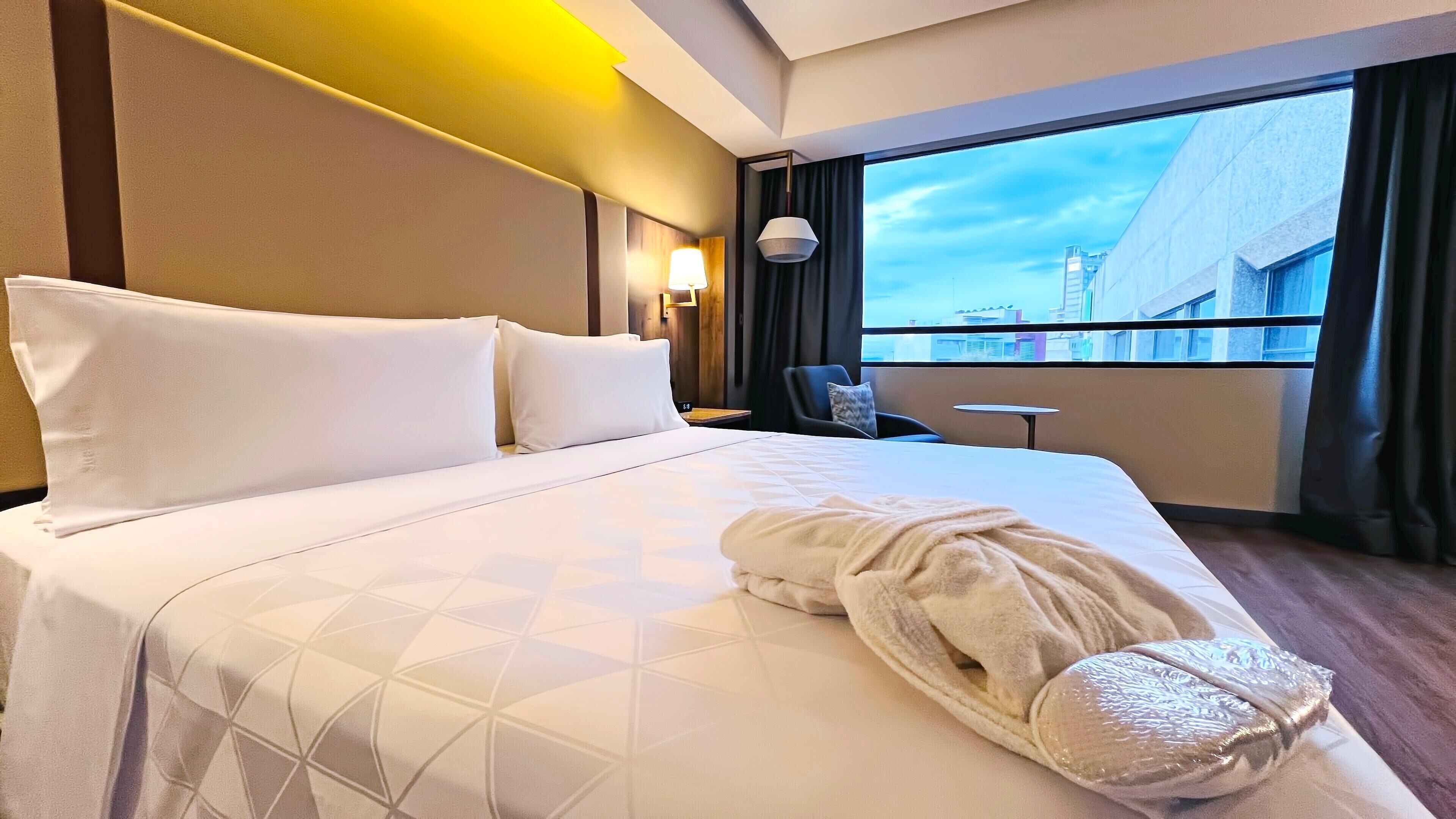 Habitación de hotel con una cama grande, una silla, una mesita y vistas a edificios a través del ventanal.