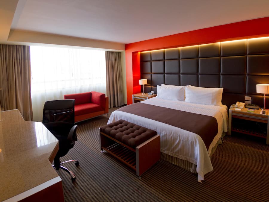 A hotel room with a bed, a desk, a chair, a small table, a telephone, and a window with curtains.