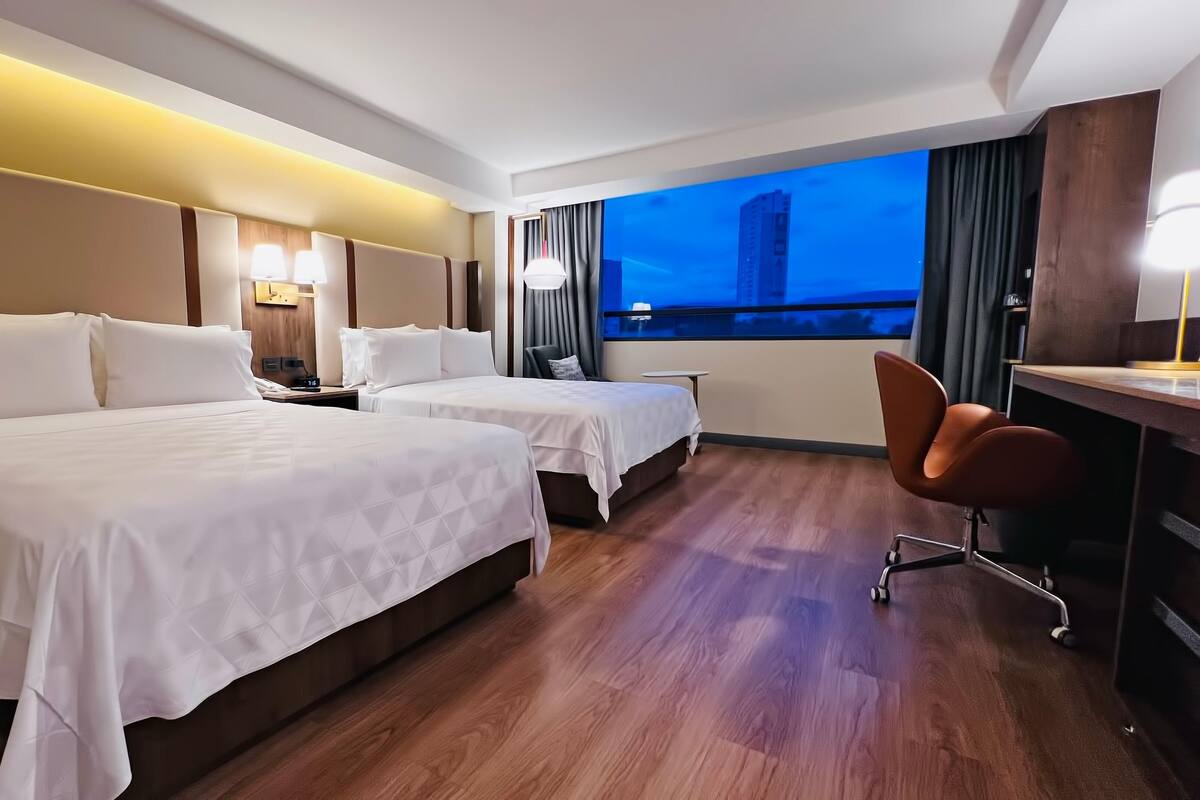 A hotel room with two beds, a desk, a chair, a nightstand, and a view of a cityscape.
