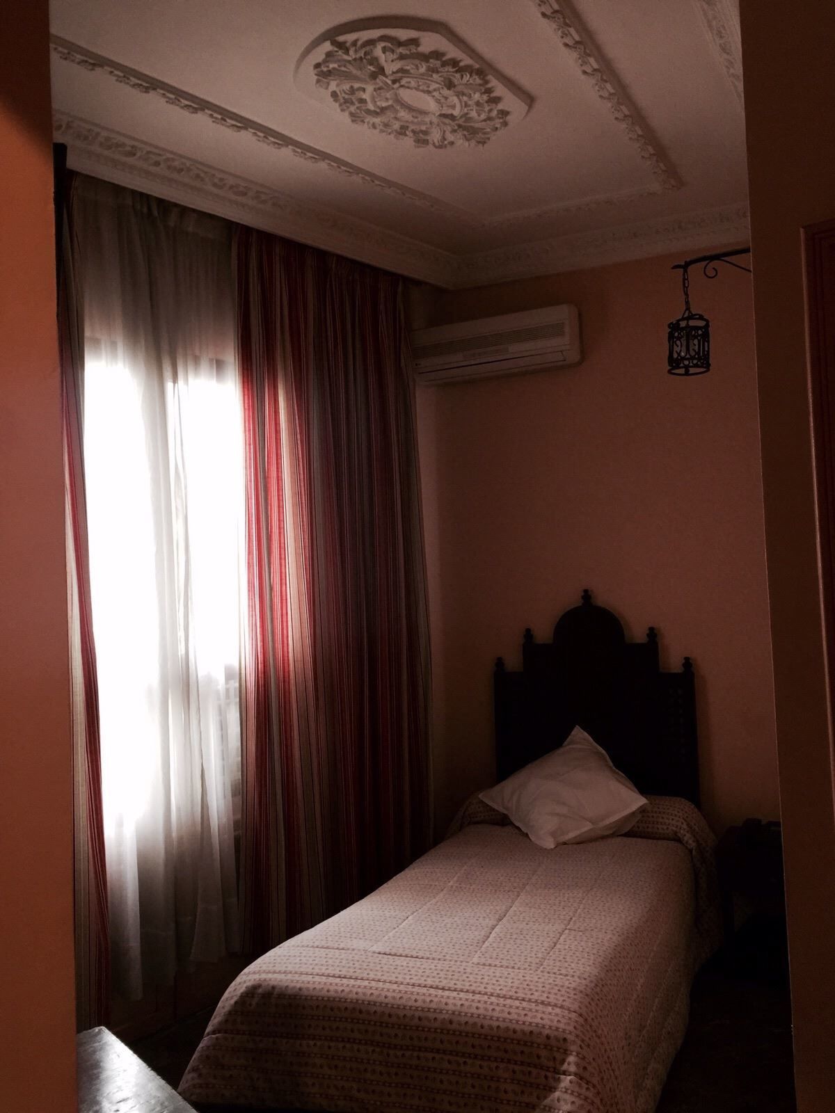 Un dormitorio con una cama individual, una ventana con cortinas, un equipo de aire acondicionado y un farol colgante.