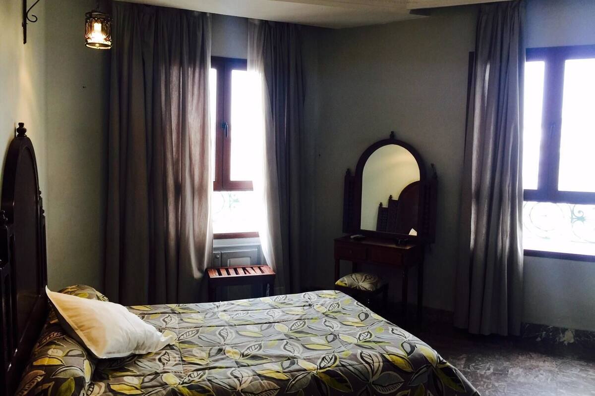 Een slaapkamer met een bed, een dressoir, een spiegel en een raam met gordijnen.