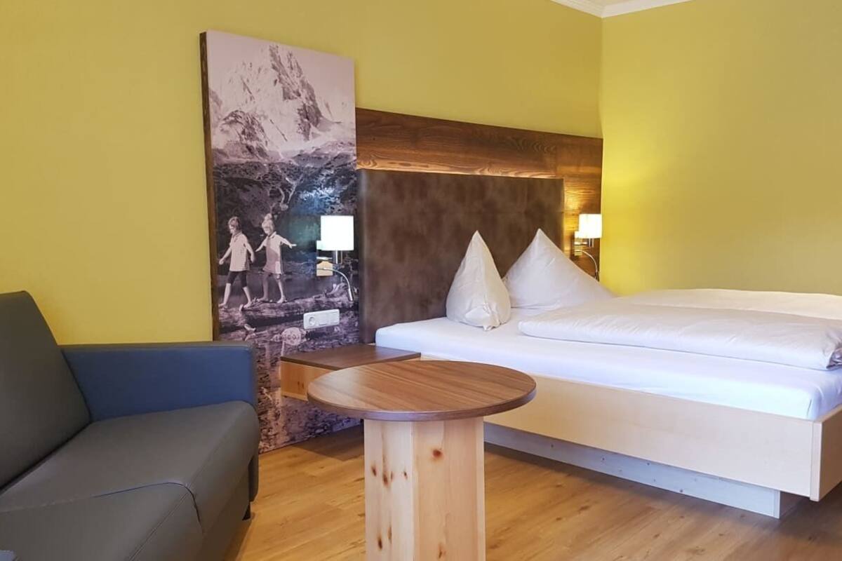 A room with a bed, a sofa, a wooden table, and a large wall painting.