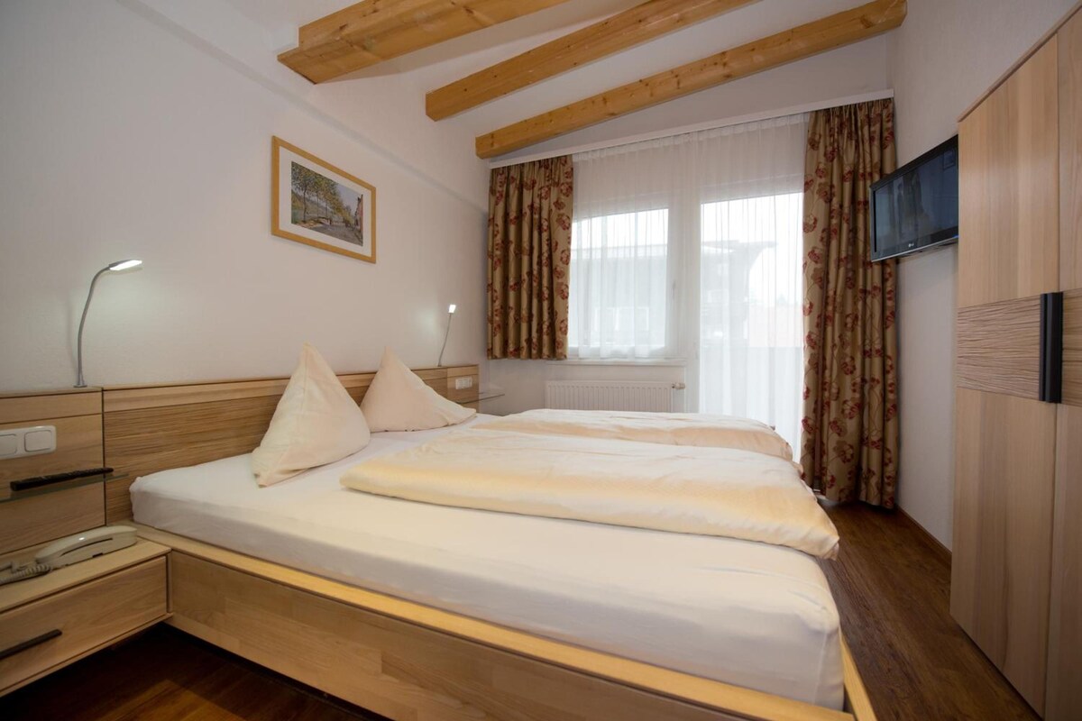 Ein Schlafzimmer mit Holzkopfteil, einem Bett mit weißer Bettwäsche, einem Nachttisch, einer Lampe, einem Bild an der Wand und einem Fenster mit Vorhängen.