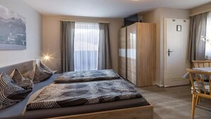 A modern bedroom with a bed, a wooden wardrobe, a desk with a chair, and a large window with curtains. - Gerlinde Feichter (Soell)