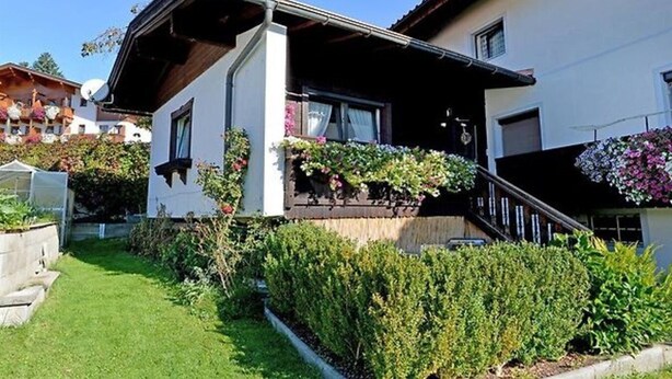 A two-story house with a white exterior, a wooden balcony, and a small garden area with various plants and flowers. - Gerlinde Feichter (Soell)
