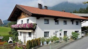 A white building with a red-tiled roof, a balcony with flowers, and a staircase leading up to the entrance. - Gerlinde Feichter (Soell)