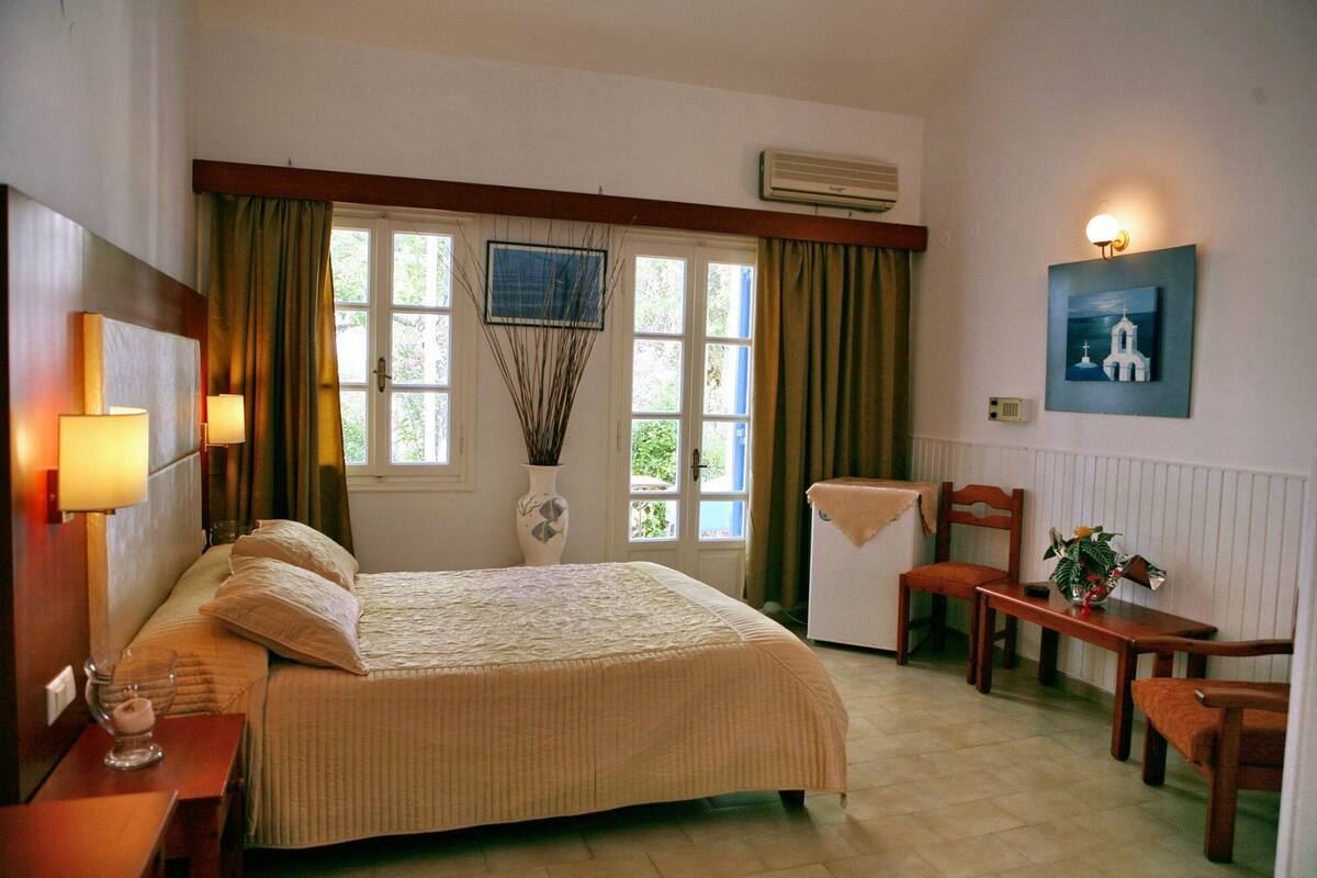 A bedroom with a bed, bedside tables, a desk, a chair, a window with curtains, and a wall-mounted air conditioner.