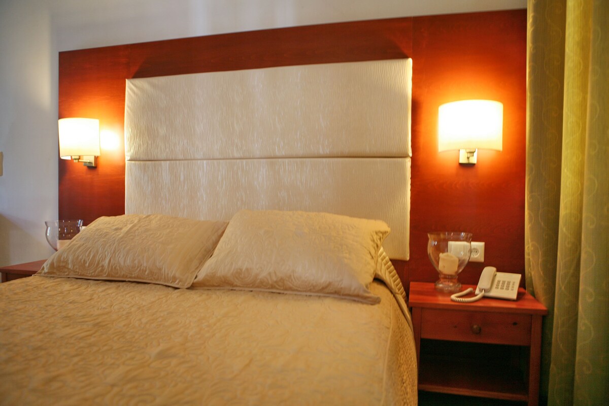 A hotel room with a large bed, two bedside lamps, a bedside table with a phone and a glass, and a wooden headboard.