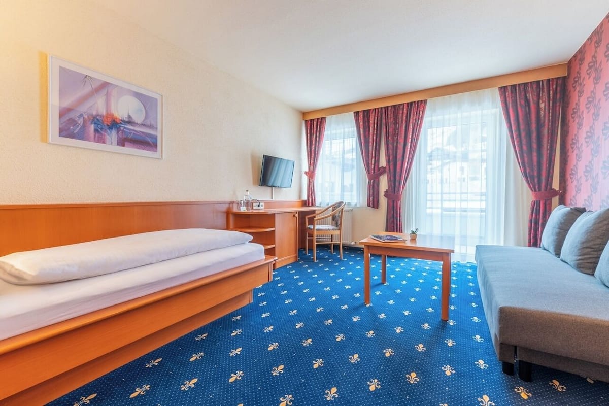 Quarto de hotel com cama, sofá, escrivaninha com cadeira, televisão e janela com cortinas.