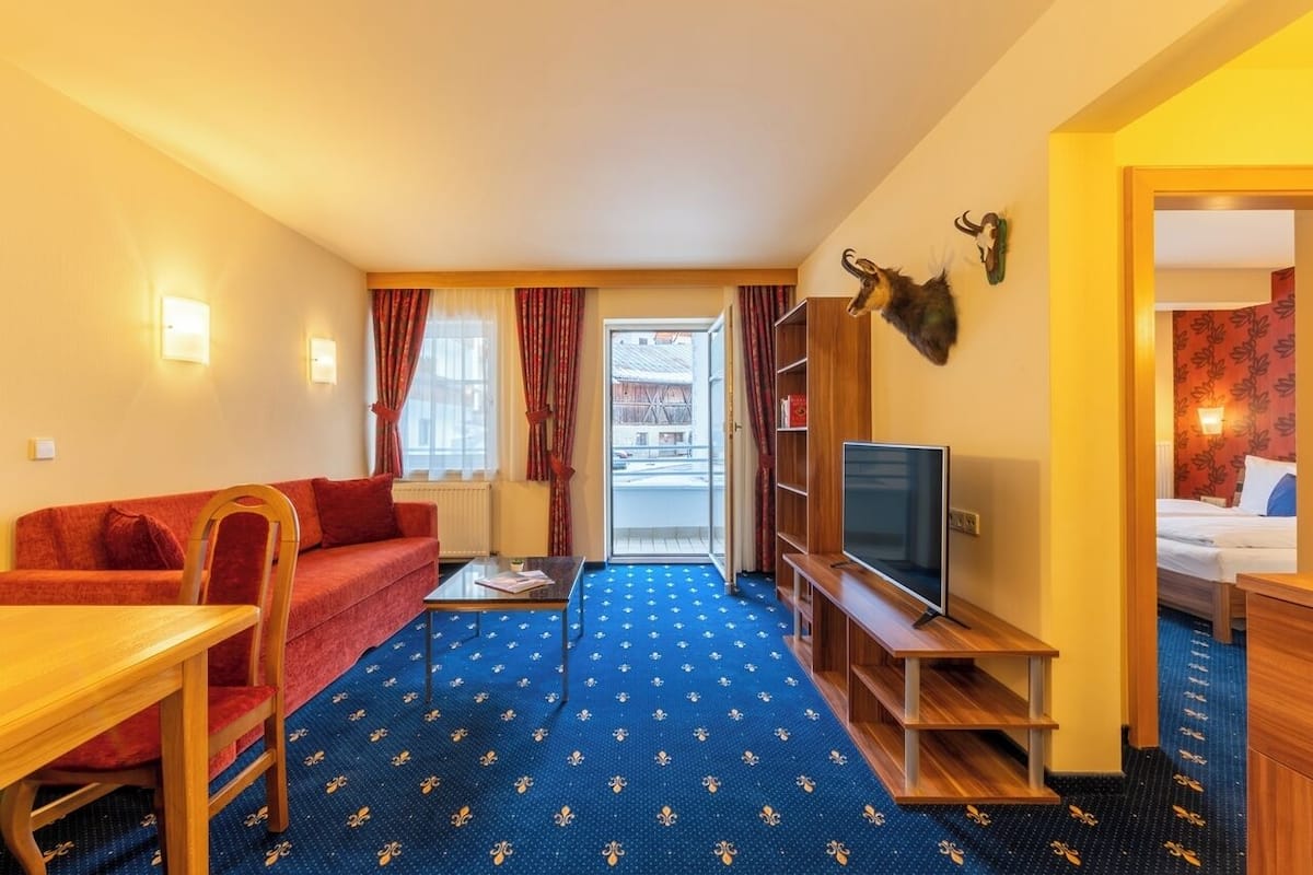 Quarto de hotel com sofá vermelho, mesa de centro de madeira, televisão em suporte de madeira e estante de livros.