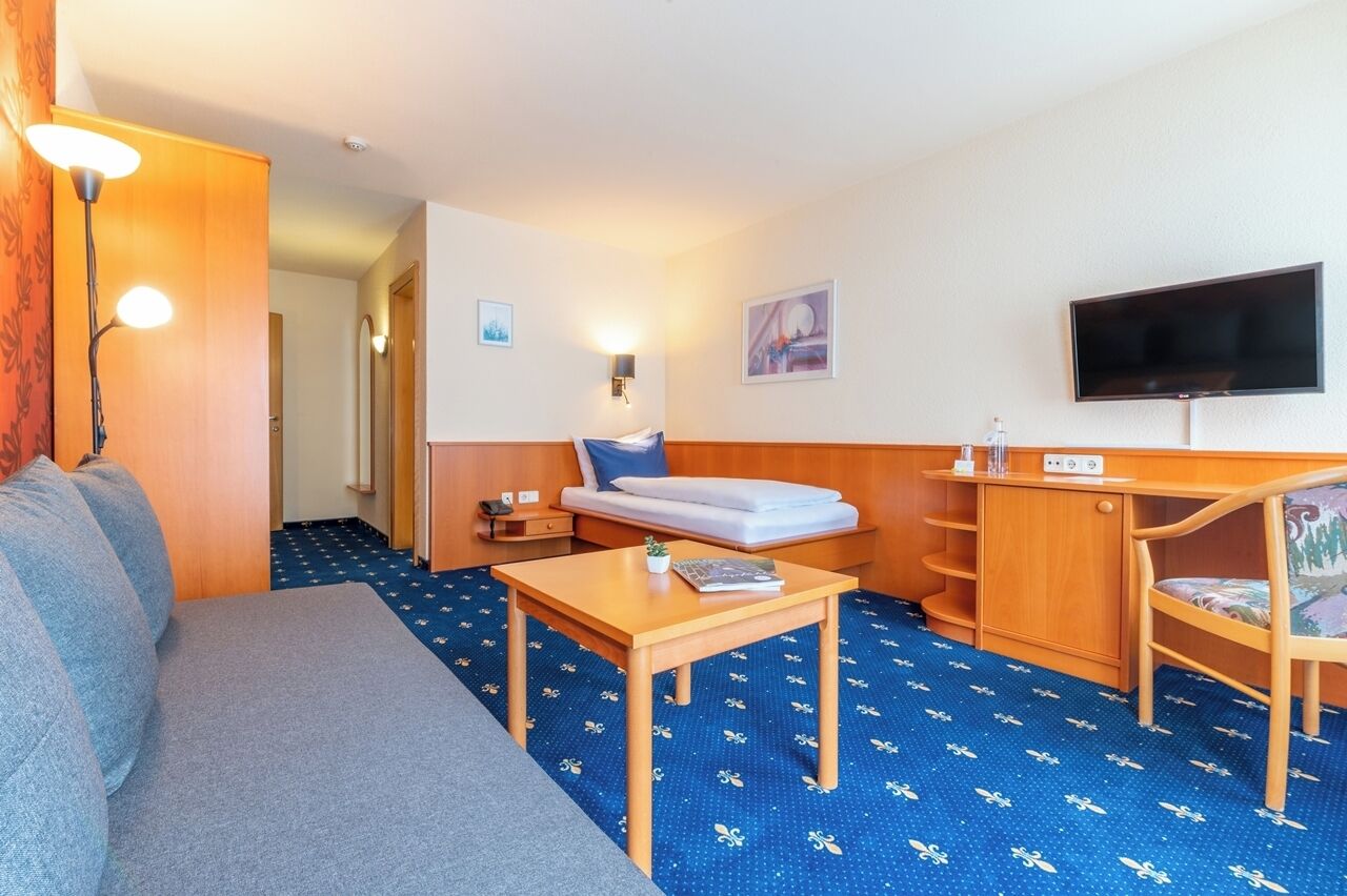 Habitación de hotel compacta con sofá, mesita pequeña, escritorio, cama, televisor y alfombra estampada.