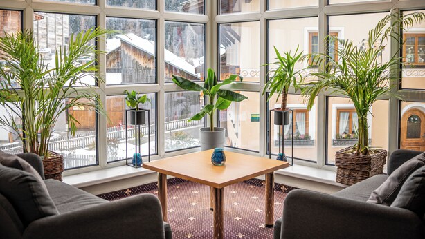 Een kamer met een groot raam dat uitkijkt op een besneeuwd berglandschap, met een tafel, stoelen en potplanten.