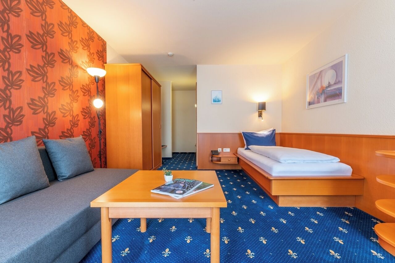 Une chambre d’hôtel avec un canapé, une petite table sur laquelle repose un magazine, un lit avec un matelas blanc, une table de chevet et une lampe fixée au mur.