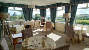 A dining area with multiple tables set for meals, large windows with curtains, and a view of the outdoors. - Trelawne Hotel (Falmouth)