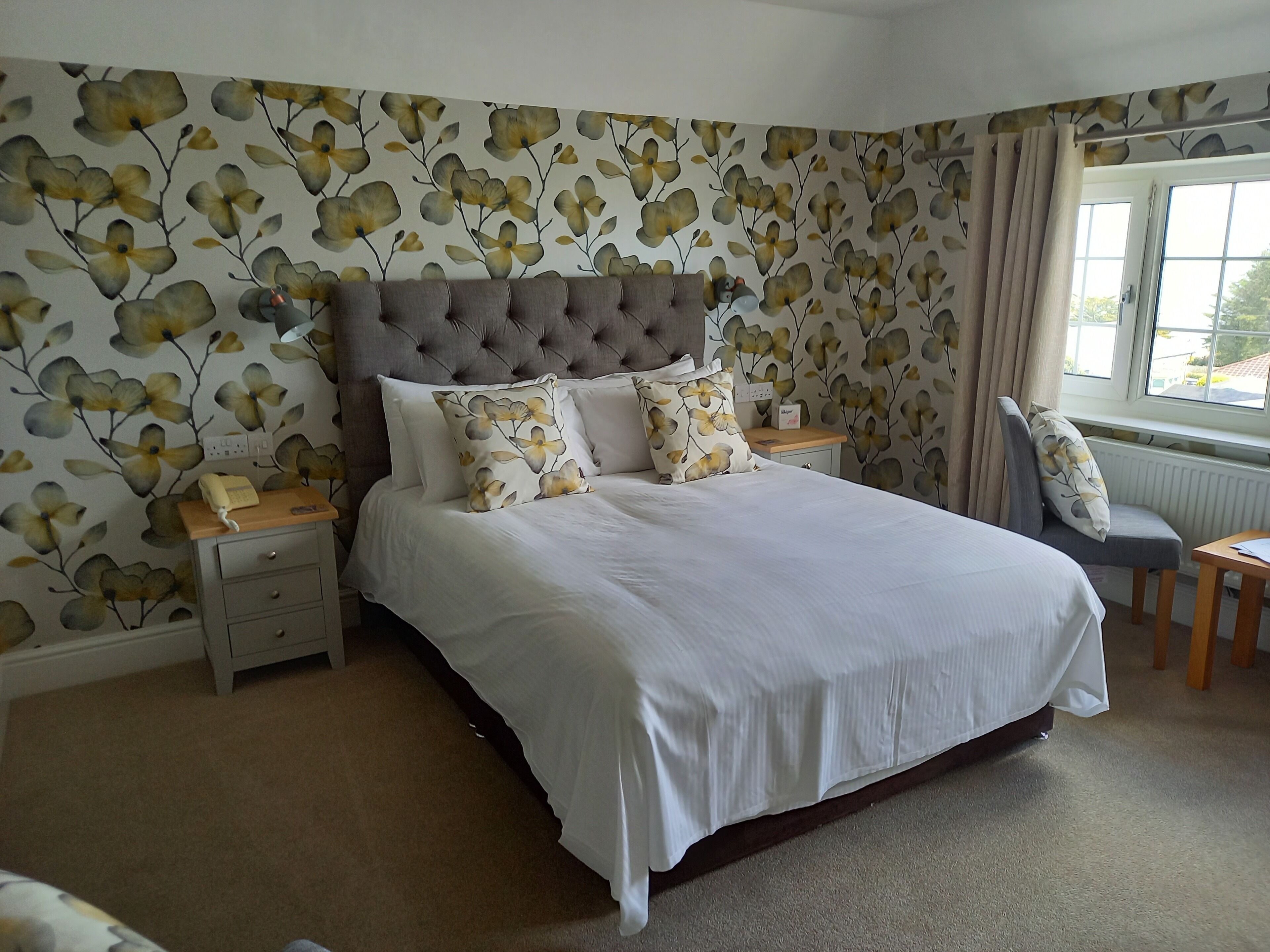 A bedroom with a large bed, a tufted headboard, floral wallpaper, a bedside table with a lamp, and a view of the outdoors through a window.