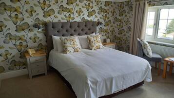 A bedroom with a large bed, a tufted headboard, floral wallpaper, a bedside table with a lamp, and a view of the outdoors through a window.