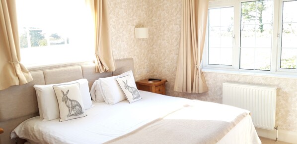 A neatly made bed with white bedding and decorative pillows, a wooden nightstand with a lamp, and a window with curtains. - Trelawne Hotel (Falmouth)