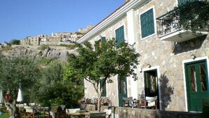A stone building with outdoor seating, a view of a hillside, and a green door. - Amfitriti (Lesvos)