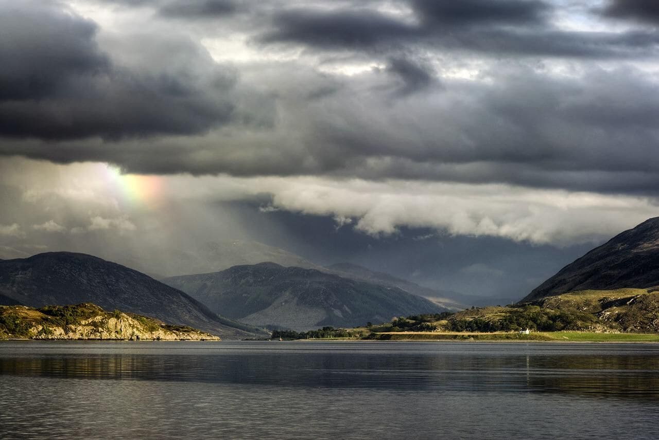 Ein See mit Bergen im Hintergrund und einem Regenbogen am Himmel.
