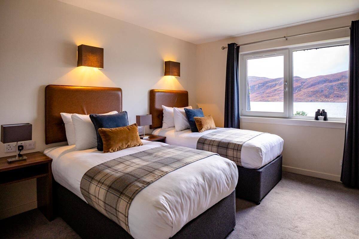 Habitación de hotel con dos camas, cada una con cabecera y lámparas de noche. Se ve una ventana con vistas a colinas y una masa de agua.
