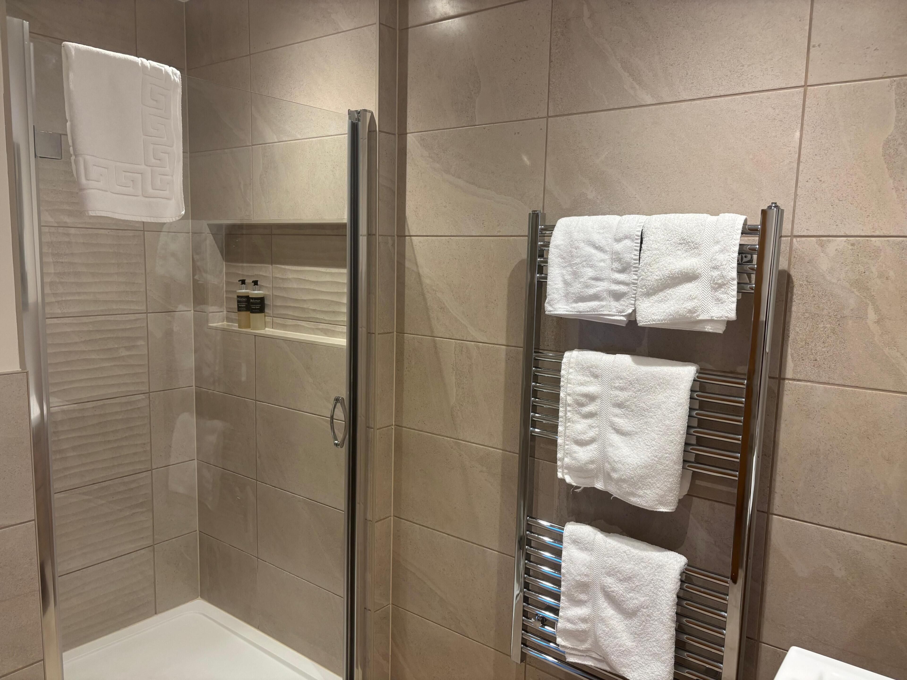 Et moderne badeværelse med et bruseafskærmning i glas, et håndklædestativ med flere håndklæder og en hylde med toiletartikler.