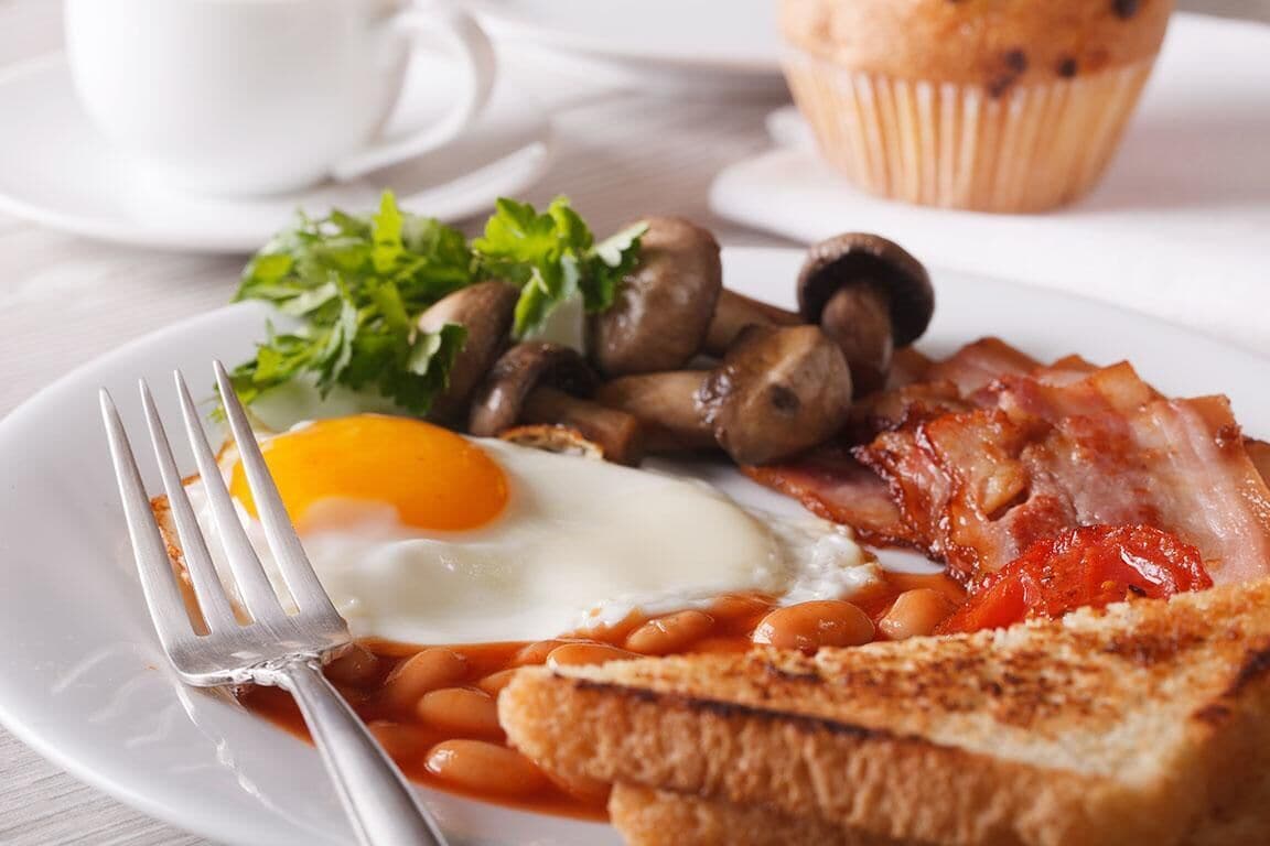 Une assiette composée d’œufs, de haricots, de champignons, de lardons, de pain grillé et d’une portion de légumes verts.
