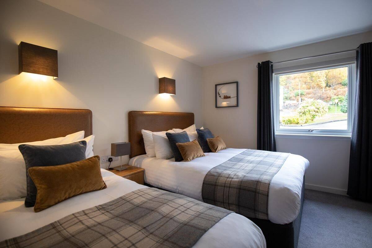 Habitación de hotel con dos camas, un ventanal con cortinas e iluminación en la pared.
