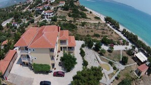 A coastal residential area with a prominent building featuring a red-tiled roof, multiple balconies, and a driveway with parked cars. - Grand Beach Hotel (Thasos)