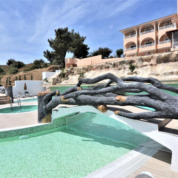 A swimming pool with a naturalistic rock feature, a white building with balconies, and a hillside with trees. - Grand Beach Hotel (Thasos)