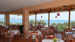 A restaurant with outdoor seating overlooking a scenic view of the sea. Tables are covered with checkered tablecloths, and there are potted plants and flowers on the tables. - Grand Beach Hotel (Thasos)