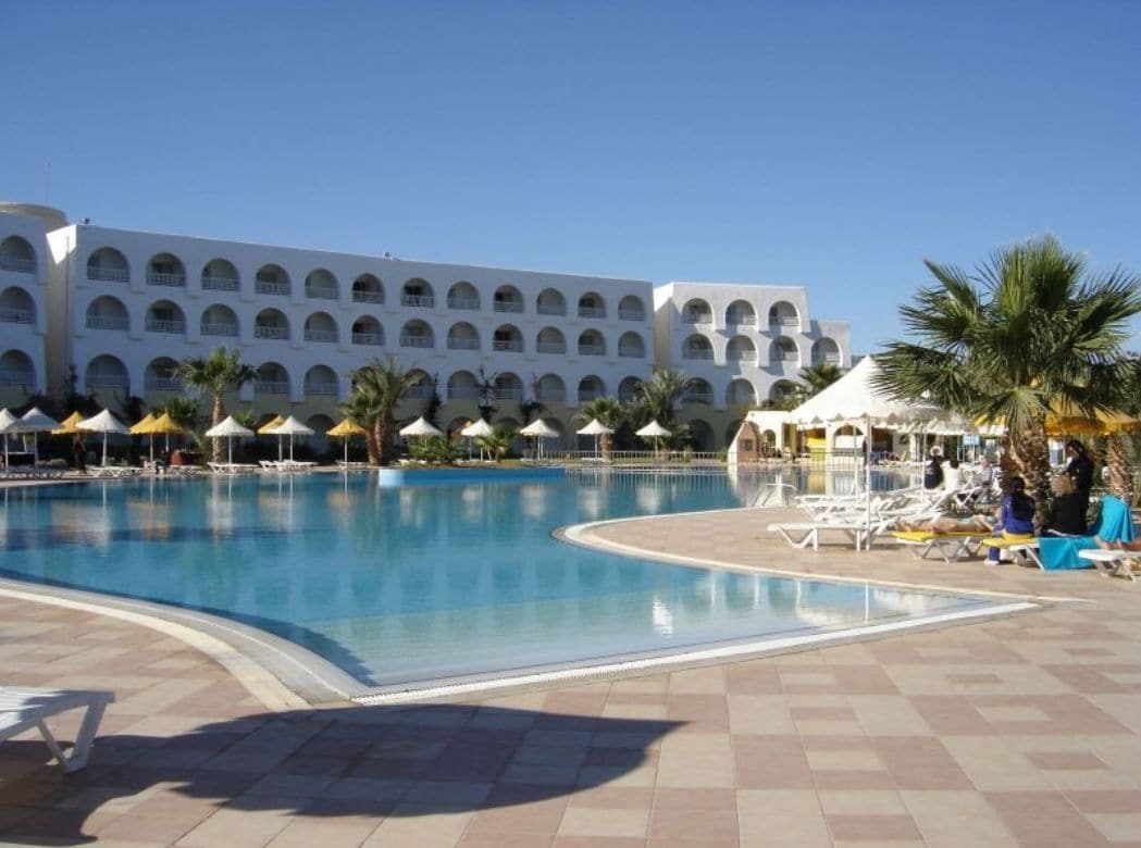 A swimming pool with lounge chairs and umbrellas, a large building with multiple balconies, and palm trees.