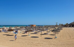 A beach with sun loungers and thatched umbrellas, a clear sky, and a building in the background. - Sidi Mansour Resort & Spa (Djerba Midun)