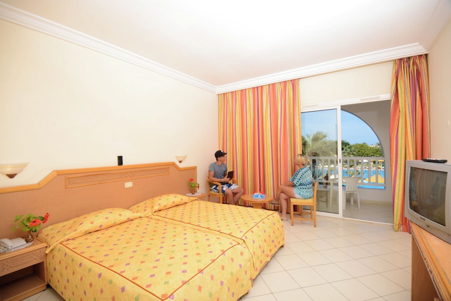 Ein Hotelzimmer mit Bett, Fernseher, einem kleinen Tisch und einem Balkon mit Blick auf einen Pool.