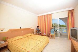 A hotel room with a bed, a television, a small table, and a balcony with a view of a pool. - Sidi Mansour Resort & Spa (Djerba Midun)