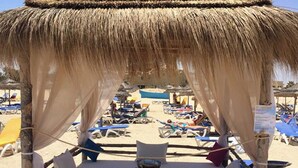 A beach cabana with a thatched roof, a small table, and cushioned seating.