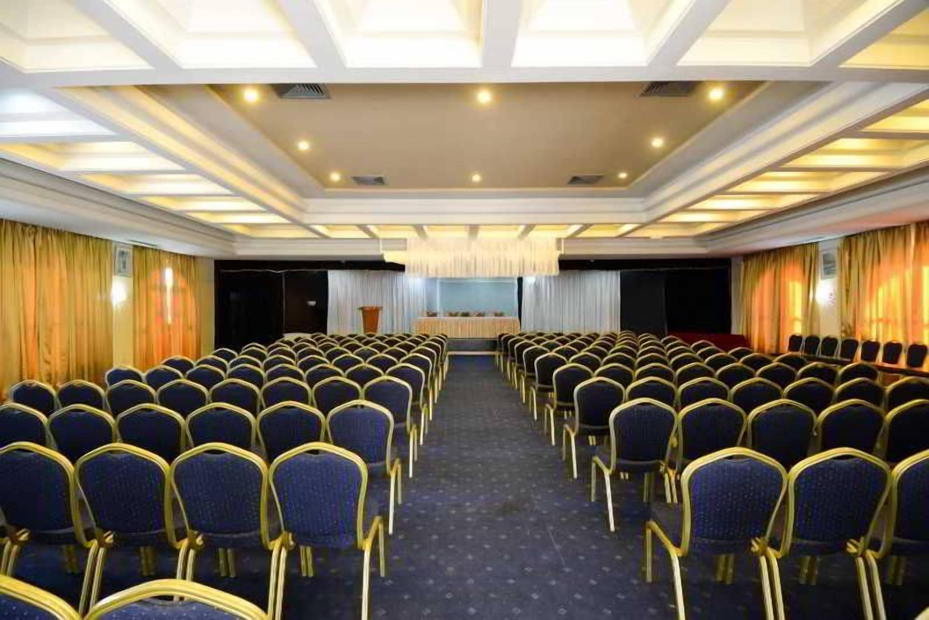 Un salón de conferencias con filas de sillas, un escenario e iluminación empotrada en el techo.