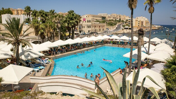Ein Swimmingpool mit Liegestühlen und Sonnenschirmen, umgeben von Palmen und Gebäuden.