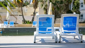 Zwei weiße Lounge-Stühle mit blauen Kissen am Pool, Palmen und ein Gebäude im Hintergrund.