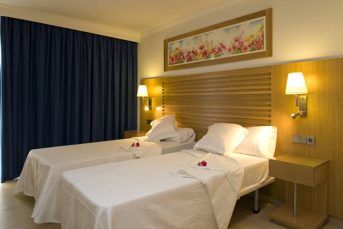 A hotel room with two beds, a painting above the headboard, and a nightstand with a lamp.