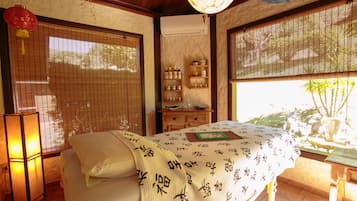 A traditional Japanese-style room with tatami flooring, a bed with a white bedspread featuring calligraphy, a wooden cabinet with bottles, and a sliding door with a view of greenery outside.