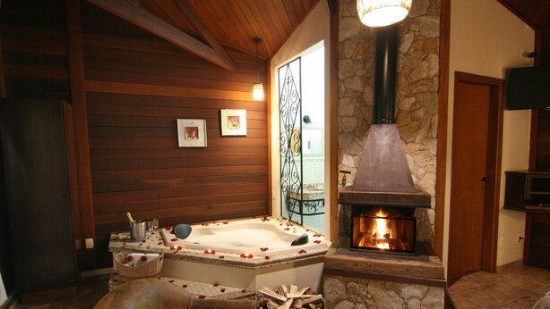 A cozy room with a stone fireplace, a wooden ceiling, a large bathtub with rose petals, and a stone wall. - Pousada Varanda das Colinas (Monte Verde)