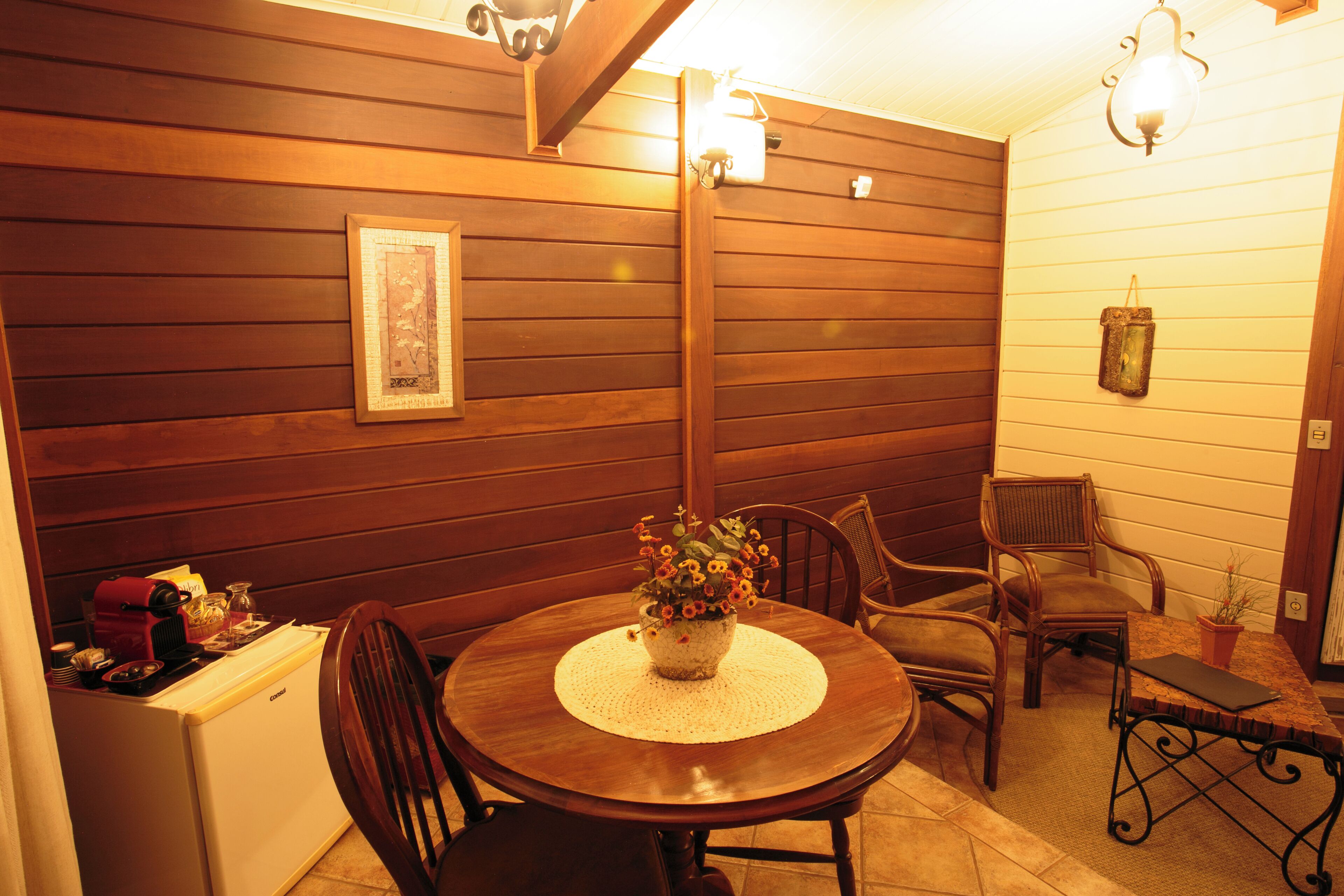 A wooden-paneled room with a dining table, chairs, a small refrigerator, and a wall-mounted light fixture.