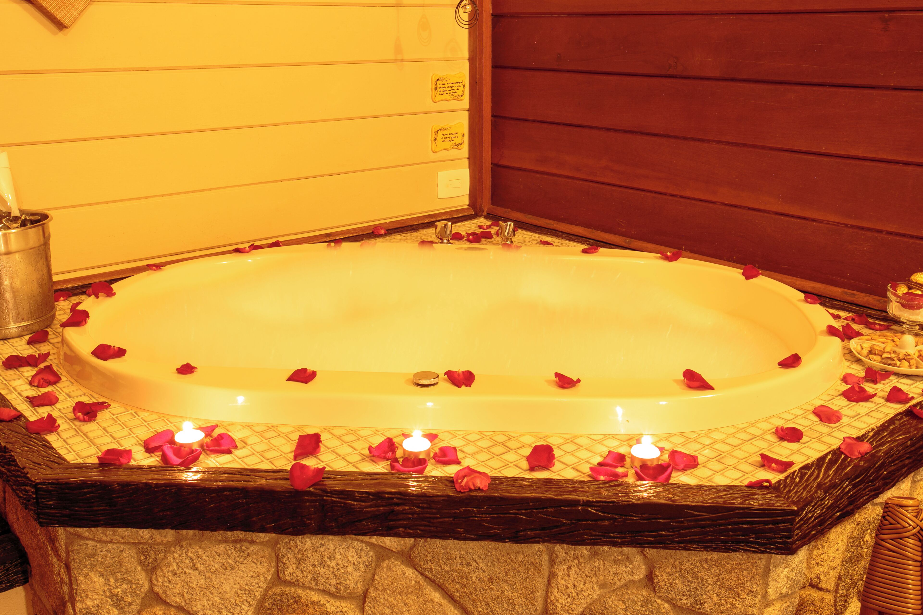 A large oval-shaped bathtub filled with rose petals and candles, surrounded by stone walls and wooden paneling.
