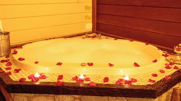 A large oval-shaped bathtub filled with rose petals and candles, surrounded by stone walls and wooden paneling.